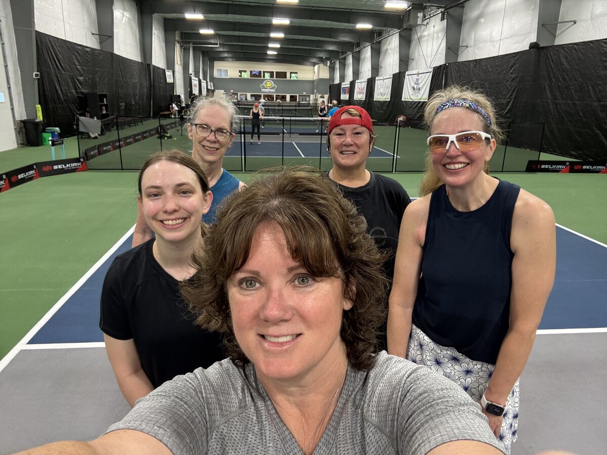Selfie of a PickleME207 coach with four clinic players smiling on an indoor pickleball court, with more courts in play behind them.