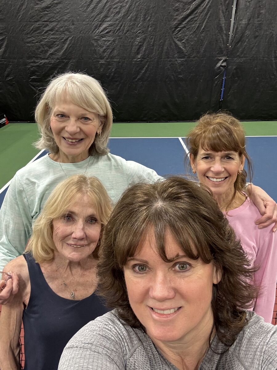 Close selfie of a PickleME207 coach with three clinic players leaning in together beside an indoor court.