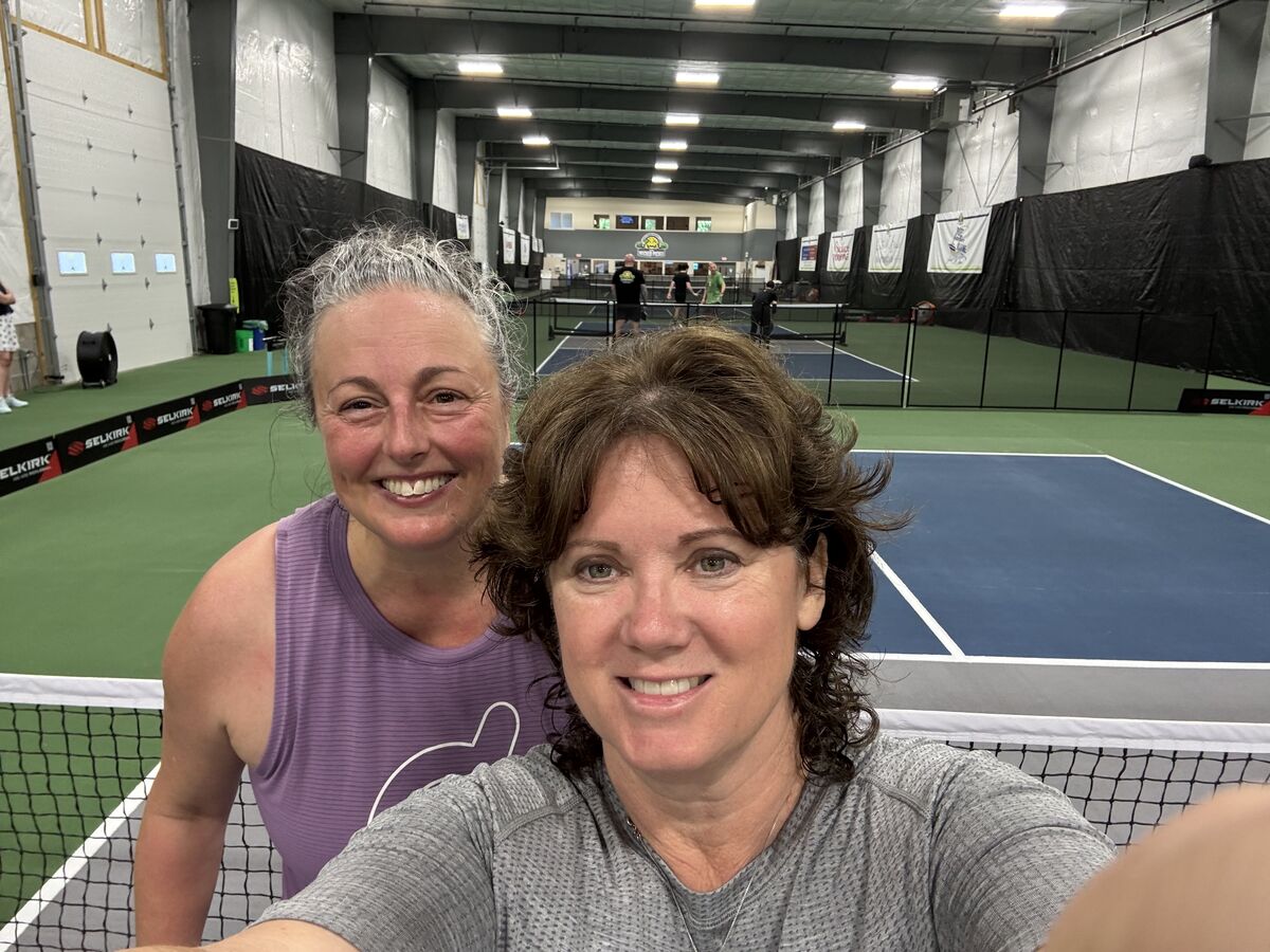 Two players grinning in a selfie taken at the net of an indoor pickleball court after a match.