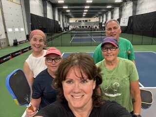 Group selfie of a PickleME207 coach and five clinic players, paddles in hand, on an indoor pickleball court.