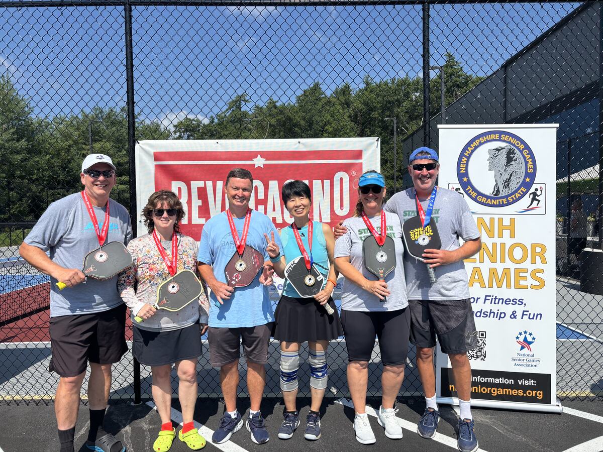 Six players holding paddles and wearing medals on an outdoor court in front of a New Hampshire Senior Games banner.